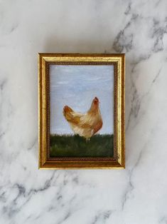 a painting of a chicken in a frame on a white marble surface with gold trim