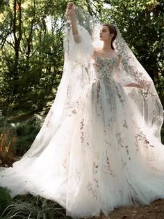 a woman wearing a wedding dress and veil standing in the woods with her arms outstretched