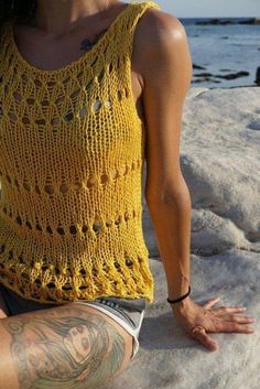 a woman sitting on top of a rock next to the ocean wearing a yellow crocheted tank top