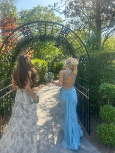 two women in dresses are walking down a path through an archway with trees and bushes on either side