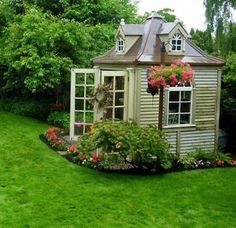 a small garden shed with flowers in the front and on the side, surrounded by lush green grass