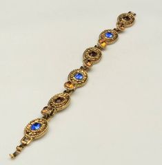 -Vintage Hollycraft gold tone multicolor rhinestone bracelet-Marked: HOLLYCRAFT, on back of each side of clasp piece-Great vintage condition: some metal rub on back side of bracelet and inside clasp, a couple of gems are unevenly glued into settings-Fold over clasp, holds tight-Blue and dark red rhinestones: approx 7.12mm x 5.45mm, oval-Amber rhinestones: approx 6.47mm, round-Wearable length: 7 3/8"-Width: a hair over .5"-Depth: .25"-Weight: 25.5g-Add this vintage Hollycraft bracelet to your jew Vintage Jeweled Gold Bracelets, Antique Jeweled Gold Bracelets, Vintage Gold Jeweled Bracelets, Vintage Gold Jeweled Crystal Bracelet, Vintage Gold Crystal Bracelet, Jeweled Gold Brass Bracelet, Gold Jeweled Brass Bracelet, Gold Bracelets With Stone Setting For Party, Vintage Gold Crystal Bracelet For Formal Occasions