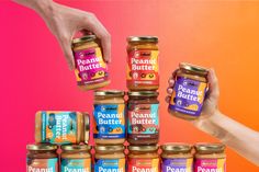 two hands reaching for peanut butter in front of a stack of jars on an orange and pink background