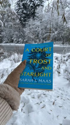 a person holding up a blue book in front of snow covered trees and bushes with the title a court of frost and starlight written on it