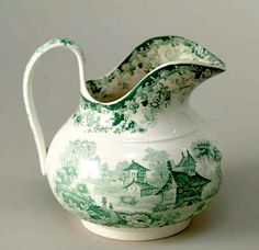 a green and white porcelain pitcher with a house on it's side, in front of a gray background