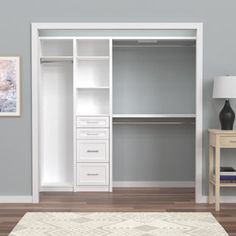 a white closet with shelves and drawers next to a lamp on a table in front of a gray wall