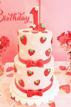 a three tiered white cake with red bows and hearts on the top is sitting on a table in front of pink flowers