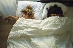 two women laying in bed with white sheets and blankets on them, one has her head under the covers
