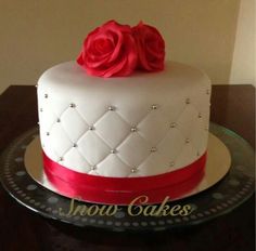 a white cake with red ribbon and two roses on top is sitting on a table