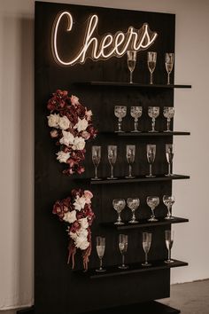 a black shelf with wine glasses and flowers on it next to a neon sign that says cheers