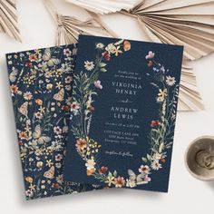 two blue wedding cards with floral designs on them next to a silver cup and fan