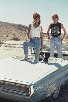 two women standing on top of an old car in the middle of nowhere, with mountains in the background