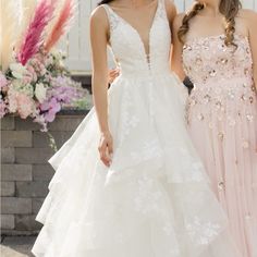 two beautiful women standing next to each other in front of a white fence and flowers