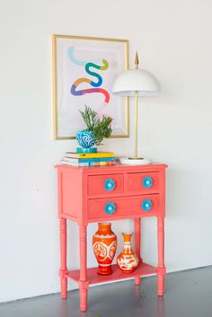 a pink table with two orange vases on it and a framed painting above it