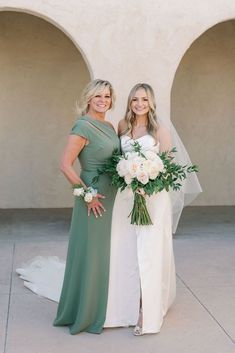 two women standing next to each other in front of a building with white flowers and greenery