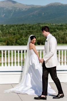 a bride and groom holding hands in front of a white fence with mountains in the background