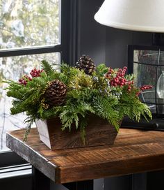 a wooden table topped with a planter filled with pine cones and greenery next to a window