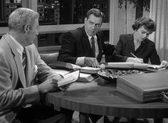 three people sitting at a table with books and papers in front of them, one man is writing