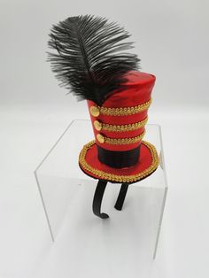 a red hat with black feathers and gold trims on top of a clear acrylic stand