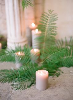 a white candle sitting on top of a stone floor next to green plants and candles