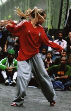 a woman in red shirt and grey pants doing a dance move on stage with people watching