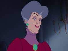 an older woman with grey hair and green earrings looks at the camera while standing in front of a chandelier