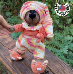 a crocheted teddy bear is posed on a wooden table with trees in the background