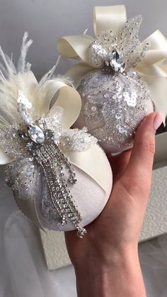 two white christmas balls with bows and sequins are held up by someone's hand