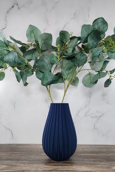 a blue vase filled with green leaves on top of a wooden table next to a marble wall