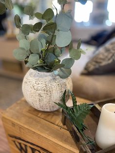 a white vase filled with greenery sitting on top of a wooden box next to a candle