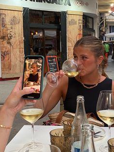 a woman sitting at a table drinking from a wine glass while looking at her cell phone