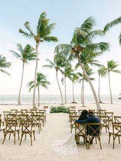 a bride and groom sitting on chairs under palm trees at the beach