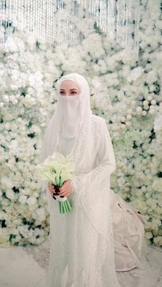 a woman dressed in white holding flowers and wearing a veil over her head is standing in front of a flower wall