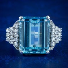 A magnificent vintage cocktail ring made in the Art Deco fashion with a colossal emerald cut aquamarine in the centre, weighing approx. 15ct with a pleasing, deep ocean blue hue.  The aqua is complemented by an audience of twenty-four sparkling brilliant cut diamonds arranged in a graduated, stepped design down each shoulder. They're 0.05ct each (approx. 1.20ct total) and have superb SI1 clarity - H colour.  The robust gallery is modelled entirely in 18ct white gold and displays the stones high Formal Light Blue Diamond Ring, Luxury Formal Blue Diamond Ring, Formal Aquamarine Diamond Ring With Brilliant Cut, Aquamarine Brilliant Cut Diamond Ring For Formal Occasions, Formal Light Blue Aquamarine Diamond Ring, Luxury Gia-certified Emerald Cut Topaz Ring, Gia Certified Blue Art Deco Jewelry, Blue Art Deco Jewelry Gia Certified, Blue Octagon Diamond Ring With Center Stone