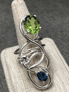 I've created this Bohemian abstract large finger ring, a true statement piece, in sterling silver, which features a 10x8mm oval Peridot, a 6mm White Topaz and an 8x6mm Blue Zircon. I make everything in 925 Sterling Silver and can Rhodium plate if you wish over the piece. I use AA+ grade gemstones in my work. The ring base is 12 gauge round sterling silver wire. No two are alike has I hand-fabricate one-of-a-kind designs. This statement ring is sturdy and I soldered many contact points and harden Modern Silver Jewelry With Peridot, Modern Silver Peridot Jewelry, Silver Fusion Ring With Accent Stones, Modern Silver Topaz Ring With Gemstone Accents, Silver Fusion Rings With Gemstone Accents, Unique Multi-stone Peridot Rings, Sterling Silver Rings With Gemstone Accents In Fusion Style, Unique Sterling Silver Ring With Gemstone Accents, Sterling Silver Rings With Gemstone Accents