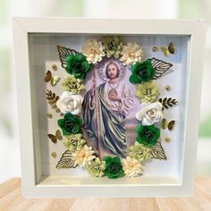 an image of jesus surrounded by flowers in a white frame on top of a wooden table