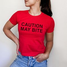 Do you bite back? Let everyone know with this "Caution: May Bite" graphic tee. Inspired by vet warning labels and your general lack of patience. Bold black lettering on a minimal design makes this the perfect piece for your grunge wardrobe, effortlessly blending sarcasm with street style. Whether you're channeling your sarcastic humor or just having a grumpy day, this statement tee is the perfect warning label for the world.  * Bold black text  * Perfect for those who like to come to the functio Edgy Fitted Slogan T-shirt, Edgy Fitted T-shirt With Slogan, Red Punk Tops With Letter Print, Punk Red Tops With Letter Print, Punk Style Red Tops With Letter Print, Red Letter Print Edgy Tops, Red Edgy Tops With Letter Print, Edgy Red Tops With Letter Print, Band Merch Crew Neck Top With Funny Text