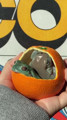 a person holding an orange with a small toy in it's mouth and the face of a frog inside