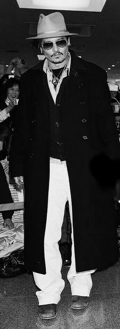 black and white photograph of a man wearing a coat, hat and sunglasses at an airport