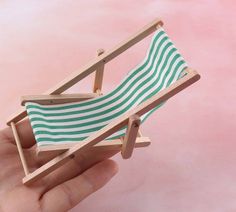 a hand holding a miniature wooden chair with green and white striped fabric on it, against a pink background