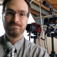 a man wearing glasses and a tie standing in front of a camera set up for filming