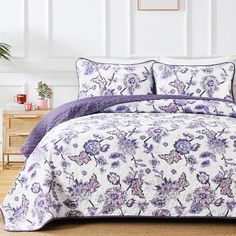 a bed covered in purple and white floral comforter sets with pillows on top of it