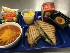 a blue tray topped with a sandwich and bowl of soup next to a container of fruit