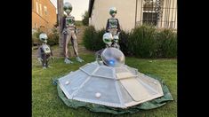 three silver alien statues sitting on top of a lush green field in front of a house