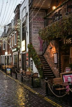 a cobblestone street lined with buildings and tables under string lights on a rainy day