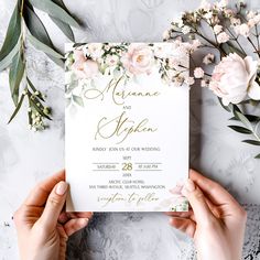 two hands holding up a wedding card with flowers and greenery around it on a table