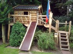 a wooden play structure with a slide in the grass next to trees and bushes on either side