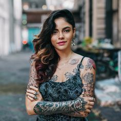 a woman with tattoos on her arms and chest standing in front of a building, looking at the camera