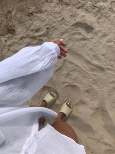 a person sitting on the beach with their feet in the sand and wearing white clothing