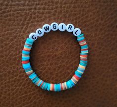 a beaded bracelet with the word cowgirl written in white and blue beads sits on a brown leather surface